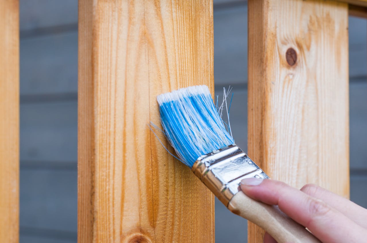 Prestations A hand painting a wooden fence plank with a blue paintbrush outdoors. Perfect for DIY and renovation themes.